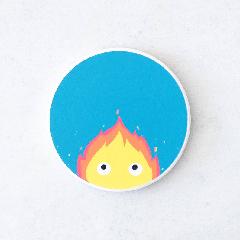 Puede incluir: Un posavasos redondo con un dise&ntilde;o de demonio de fuego de dibujos animados. El demonio tiene llamas amarillas con detalles en naranja y rojo, y dos ojos negros. El fondo es azul brillante, con un borde blanco.