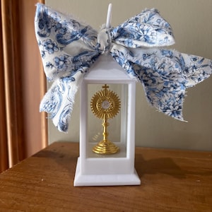 May include: A white lantern with a gold religious symbol inside, topped with a large bow. The bow features a blue floral pattern on a white background. The lantern is sitting on a wooden surface.