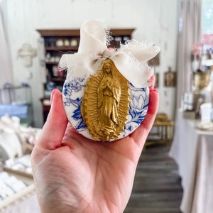 Może przedstawiać: Okrągła ozdoba ze złotą figurą religijną na białym i niebieskim kwiatowym tle, zawiązana kremową wstążką. Projekt ozdoby przypomina ikonografię religijną. Ogólnym motywem jest dekoracja religijna.