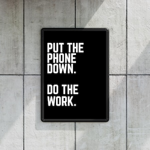 Könnte beinhalten: Ein schwarzer gerahmter Poster mit dem Text "PUT THE PHONE DOWN. DO THE WORK." in fetten weißen Buchstaben. Das Poster ist an einer grauen Betonwand angebracht. Das Bild ist gut beleuchtet, wobei der Text im Mittelpunkt steht.
