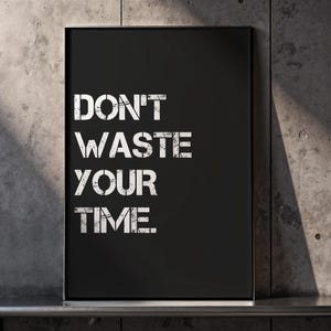 Könnte beinhalten: Schwarz gerahmter Kunstdruck mit dem Text "DON'T WASTE YOUR TIME" in weißer, abgenutzter Schrift. Das Kunstwerk wird vor einer strukturierten grauen Betonwand präsentiert, was eine moderne und motivierende Ästhetik schafft.