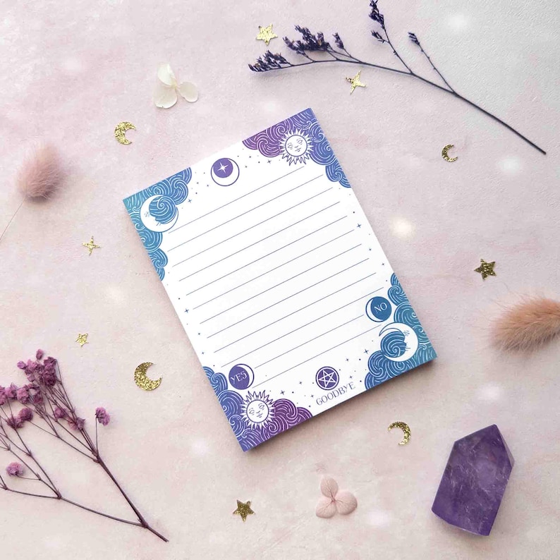 This photo is an overhead view of a notepad with a spirit board, jewel toned gradient design and college ruled lines. The design features hand drawn, swirling clouds in each corner with a moon or sun with a face, classic spirit board text like yes, no, goodbye, and a pentagram and sparkles.