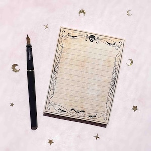 This image shows a college ruled lined 3.7 by 4.9 inch notepad with faux antiqued paper and a symmetrical vintage-inspired design that features a skull, ornate flourishes and a spiderweb border.