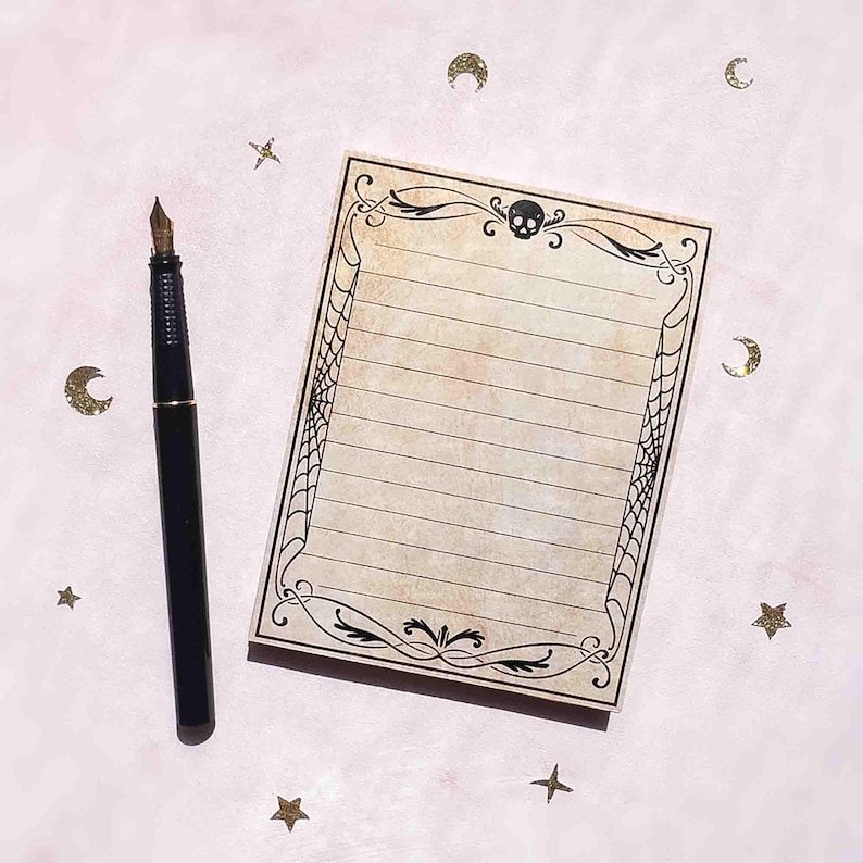 This image shows a college ruled lined 3.7 by 4.9 inch notepad with faux antiqued paper and a symmetrical vintage-inspired design that features a skull, ornate flourishes and a spiderweb border.
