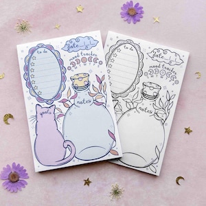 This photo shows an overhead view of two small weekly planner notepads with the same design. One has blue outlines and pastel colors, and the other is black and white. The design features an illustration of a swirly cloud that has space for you to write the week's date, a celestial picture frame that has lines and chubby stars to check off your to do list, an outline of a cat that has space for you to write out your goals, a moon phase mood tracker, and a potion bottle to write your notes in.