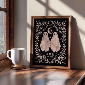 This photo shows a vertical art piece that has a black background with illustrations in taupe of two ghosts holding hands, surrounded by stars and a leaf border. A moon with a face is above the ghosts with the text Till We Are Ghosts arced above that. Below the ghosts are the couple’s initials and the year they established their relationship. The font is gothic black letter style.