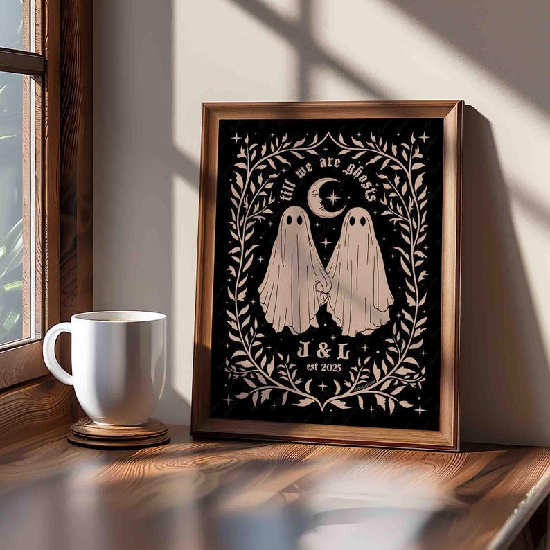 This photo shows a vertical art piece that has a black background with illustrations in taupe of two ghosts holding hands, surrounded by stars and a leaf border. A moon with a face is above the ghosts with the text Till We Are Ghosts arced above that. Below the ghosts are the couple’s initials and the year they established their relationship. The font is gothic black letter style.