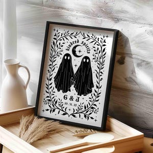 This photo shows a vertical art piece that has a white background with illustrations in black of two ghosts holding hands, surrounded by stars and a leaf border. A moon with a face is above the ghosts with the text Till We Are Ghosts arced above that. Below the ghosts are the couple’s initials and their wedding date, shown as 10.31.22. The font is gothic black letter style.