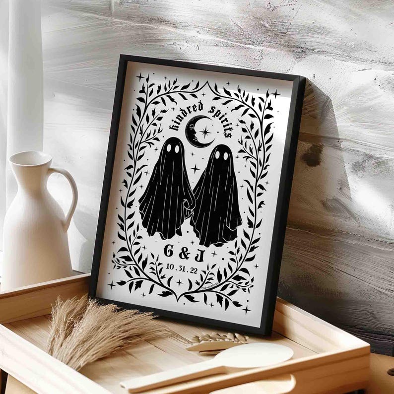This photo shows a vertical art piece that has a white background with illustrations in black of two ghosts holding hands, surrounded by stars and a leaf border. A moon with a face is above the ghosts with the text Till We Are Ghosts arced above that. Below the ghosts are the couple’s initials and their wedding date, shown as 10.31.22. The font is gothic black letter style.