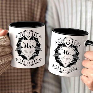 May include: Two white ceramic mugs with black handles and black rims. The mugs feature a black and white illustration of bats and a heart with the text "Mrs. Est. 2025" on one mug and "Mx. Est. 2025" on the other.