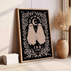 This photo shows a vertical art piece that has a black background with illustrations in taupe of two ghosts holding hands, surrounded by stars and a leaf border. A moon with a face is above the ghosts with the text Till We Are Ghosts arced above that. Below the ghosts are the couple’s initials and the year they established their relationship. The font is gothic black letter style.
