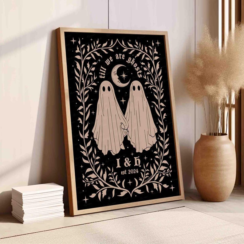 This photo shows a vertical art piece that has a black background with illustrations in taupe of two ghosts holding hands, surrounded by stars and a leaf border. A moon with a face is above the ghosts with the text Till We Are Ghosts arced above that. Below the ghosts are the couple’s initials and the year they established their relationship. The font is gothic black letter style.