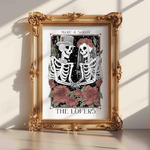 May include: A framed print featuring two skeletons dressed as a bride and groom, with the text MARC & SARAH and THE LOVERS. The skeletons are surrounded by pink roses and green leaves against a black background, all inside an ornate gold frame.