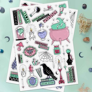 This photo showcases the second sheet of this two sheet set which has potions, candles, crystals, spellbooks, a moth in a cloche, mushrooms, sparkles, moth wall art, and a cauldron.