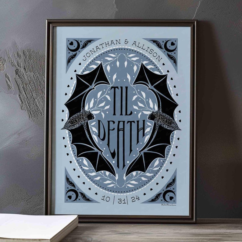 This photo shows the dusty blue and black version of this art. The art features two bats. Their wings form the shape of a heart with the words til death framed the center of the art. Crescent moons, botanical illustrations and small hearts surround the bats. The couple’s first names and anniversary date are arched on the top and bottom on the art in a folk-style font