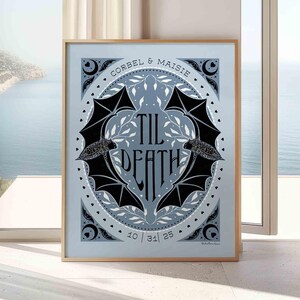This photo shows a framed poster print leaning against a wall. The art on the poster is misty blue, with a lighter blue as the background. Two illustrated bats are in the center, their wings forming a heart in the negative space, and the words Til Death in the center of the heart. White leaves are behind them, tiny hearts surround the bats and leaves, and in the corners are crescent moons, sparkles, hearts, and small bat wings. The couple’s names and date are at the top and bottom