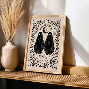 This photo shows a vertical art piece that has a beige background with illustrations in black of two ghosts holding hands, surrounded by stars and a leaf border. A moon with a face is above the ghosts with the text Till We Are Ghosts arced above that. Below the ghosts are the couple’s initials and their wedding date, shown as 10.31.25. The font is gothic black letter style.