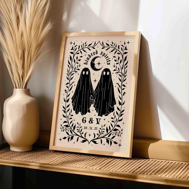 This photo shows a vertical art piece that has a beige background with illustrations in black of two ghosts holding hands, surrounded by stars and a leaf border. A moon with a face is above the ghosts with the text Till We Are Ghosts arced above that. Below the ghosts are the couple’s initials and their wedding date, shown as 10.31.25. The font is gothic black letter style.