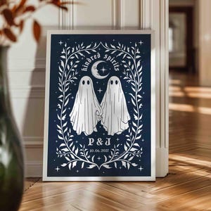This photo shows a vertical art piece that has a navy blue background with illustrations in white of two ghosts holding hands, surrounded by stars and a leaf border. A moon with a face is above the ghosts with the text Kindred Spirits arced above that. Below the ghosts are the couple’s initials and the year they established their relationship. The font is gothic black letter style.