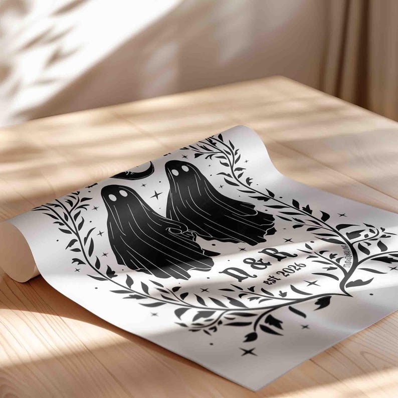 This photo shows a vertical poster partially rolled. The art has a white background with illustrations in black of two ghosts holding hands, surrounded by stars and a leaf border. A moon with a face is above the ghosts with the text Till We Are Ghosts arced above that. Below the ghosts are the couple’s initials and their wedding date, shown as est 2026. The font is gothic black letter style.