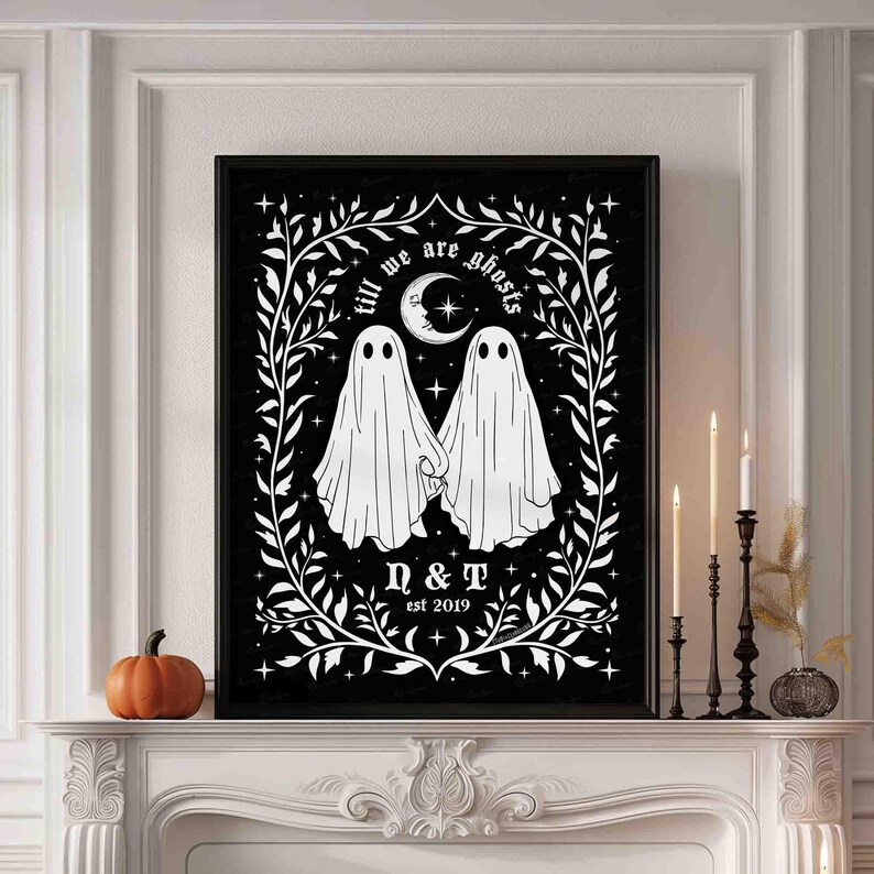This photo shows a vertical art piece that has a black background with illustrations in white of two ghosts holding hands, surrounded by stars and a leaf border. A moon with a face is above the ghosts with the text Till We Are Ghosts arced above that. Below the ghosts are the couple’s initials and the year they established their relationship. The font is gothic black letter style.