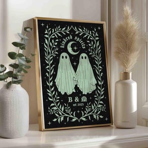 This photo shows a vertical art piece that has a black background with illustrations in sage green of two ghosts holding hands, surrounded by stars and a leaf border. A moon with a face is above the ghosts with the text Till We Are Ghosts arced above that. Below the ghosts are the couple’s initials and the year they established their relationship. The font is gothic black letter style.