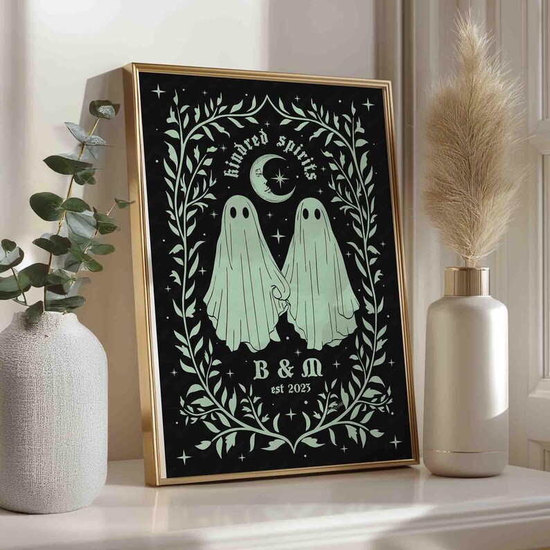 This photo shows a vertical art piece that has a black background with illustrations in sage green of two ghosts holding hands, surrounded by stars and a leaf border. A moon with a face is above the ghosts with the text Till We Are Ghosts arced above that. Below the ghosts are the couple’s initials and the year they established their relationship. The font is gothic black letter style.