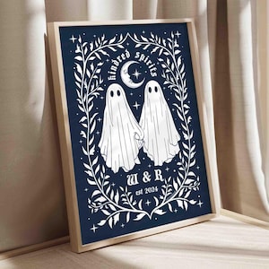 This photo shows a vertical art piece that has a navy blue background with illustrations in white of two ghosts holding hands, surrounded by stars and a leaf border. A moon with a face is above the ghosts with the text Till We Are Ghosts arced above that. Below the ghosts are the couple’s initials and the year they established their relationship. The font is gothic black letter style.