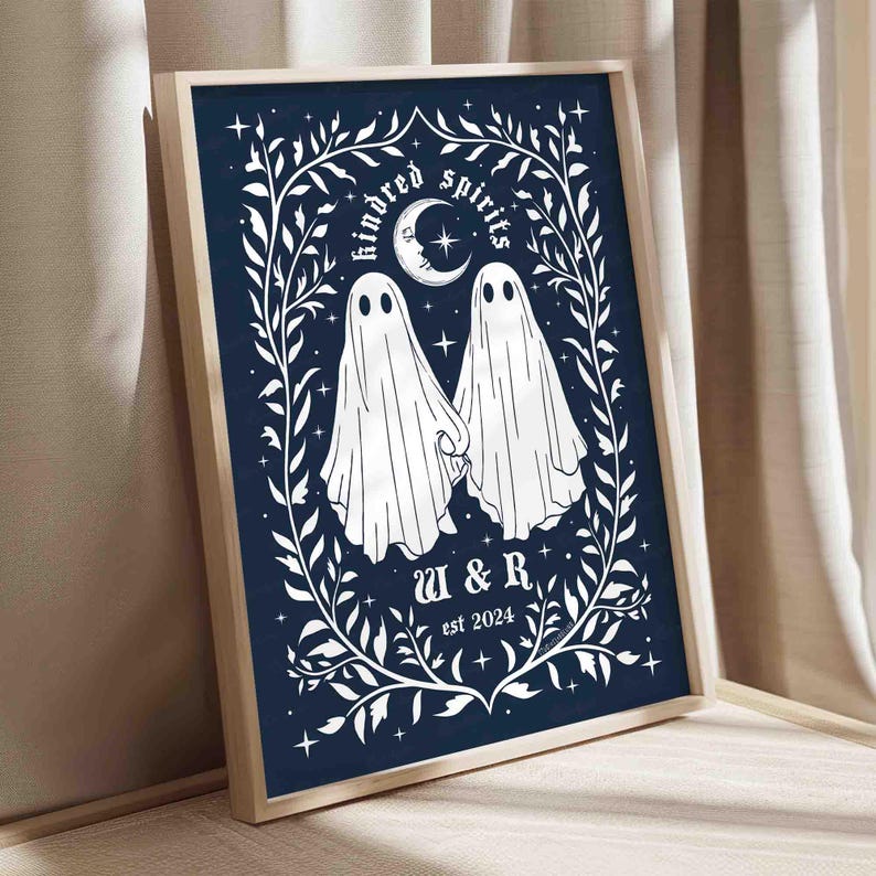 This photo shows a vertical art piece that has a navy blue background with illustrations in white of two ghosts holding hands, surrounded by stars and a leaf border. A moon with a face is above the ghosts with the text Till We Are Ghosts arced above that. Below the ghosts are the couple’s initials and the year they established their relationship. The font is gothic black letter style.
