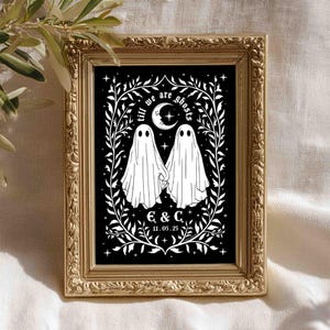 This photo shows a vertical art piece that has a black background with illustrations in white of two ghosts holding hands, surrounded by stars and a leaf border. A moon with a face is above the ghosts with the text Till We Are Ghosts arced above that. Below the ghosts are the couple’s initials and the year they established their relationship. The font is gothic black letter style.
