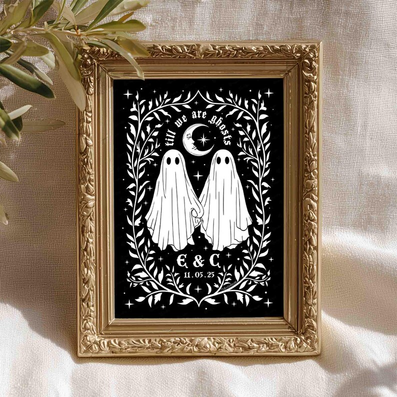 This photo shows a vertical art piece that has a black background with illustrations in white of two ghosts holding hands, surrounded by stars and a leaf border. A moon with a face is above the ghosts with the text Till We Are Ghosts arced above that. Below the ghosts are the couple’s initials and the year they established their relationship. The font is gothic black letter style.