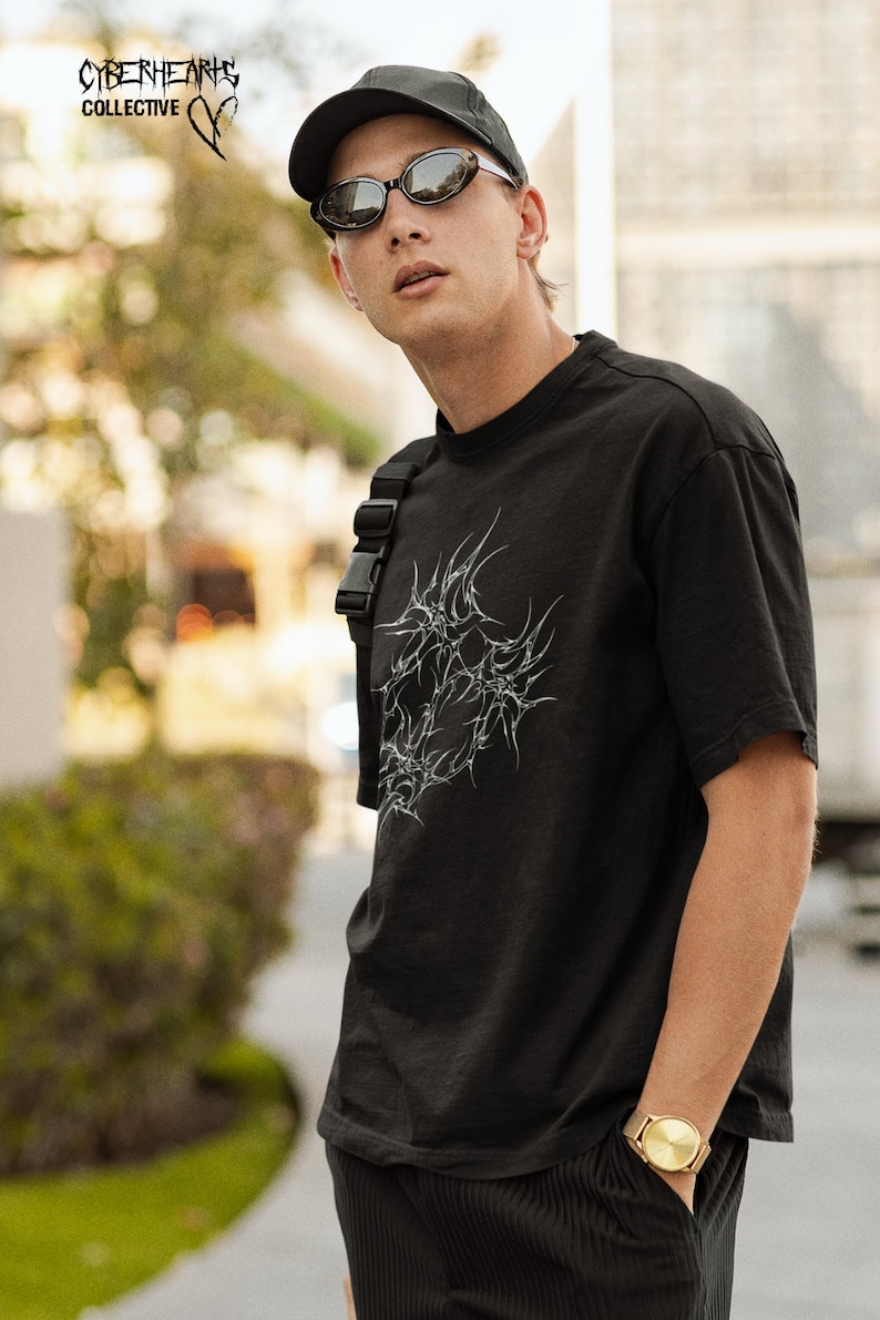 May include: A person wearing a black t-shirt with a white graphic design, black pants, a black baseball cap, and sunglasses. The t-shirt has the text "CYBERHEARTS COLLECTIVE" on the front. The person is wearing a gold watch on their left wrist.