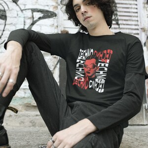 May include: A person wearing a black long-sleeved shirt with a red and white graphic print of a person wearing sunglasses and the word "techno" repeated multiple times. The person is sitting on a stone surface.