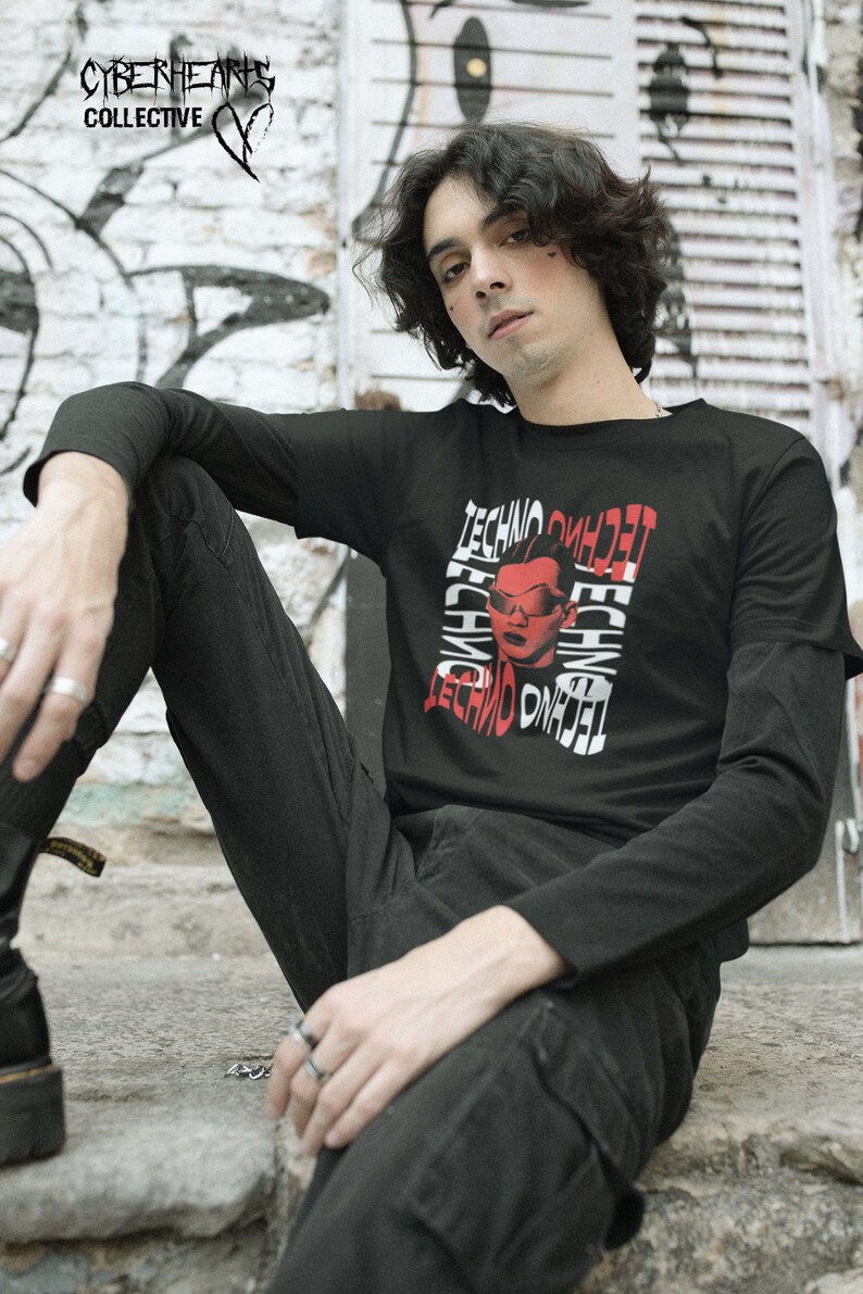 May include: A person wearing a black long-sleeved shirt with a red and white graphic print of a person wearing sunglasses and the word "techno" repeated multiple times. The person is sitting on a stone surface.