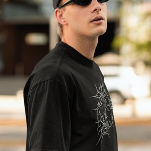 May include: A person wearing a black t-shirt with a white graphic design, a black baseball cap, and black oval sunglasses. The t-shirt has the text "CYBERHEARTS COLLECTIVE" on the top left corner.