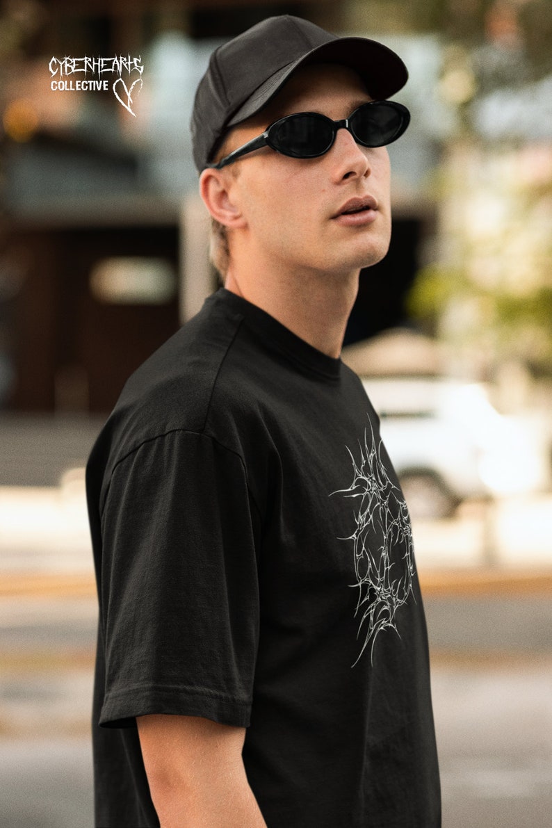 May include: A person wearing a black t-shirt with a white graphic design, a black baseball cap, and black oval sunglasses. The t-shirt has the text "CYBERHEARTS COLLECTIVE" on the top left corner.