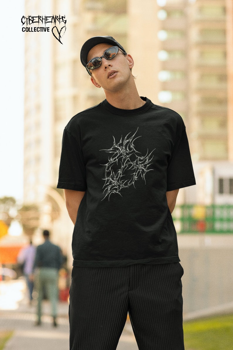 May include: A person wearing a black t-shirt with a white graphic design of a stylized heart with thorns. The t-shirt has the text "CYBERHEARTS COLLECTIVE" on the top left corner. The person is wearing a black cap and sunglasses.
