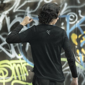May include: A person wearing a black long-sleeved shirt with a white heart design on the back. The shirt is tucked into black pants with a chain hanging from the belt loop. The person is standing in front of a graffiti-covered wall.