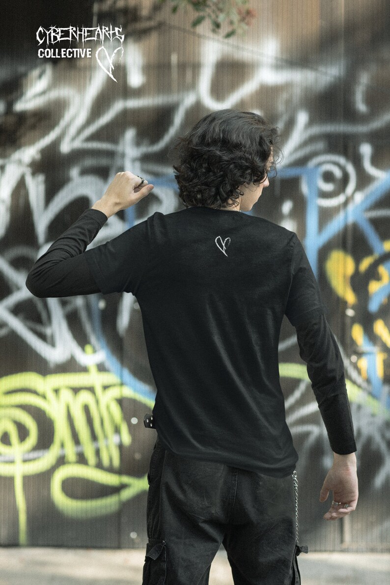 May include: A person wearing a black long-sleeved shirt with a white heart design on the back. The shirt is tucked into black pants with a chain hanging from the belt loop. The person is standing in front of a graffiti-covered wall.