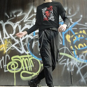 May include: A person wearing a black long-sleeved shirt with a red and white graphic print that says "Techno Omnia" and black cargo pants. The person is wearing black combat boots and a silver chain.