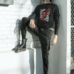 May include: A person wearing a black long-sleeved shirt with a red and white graphic print that says "Techno Omnia Vincit". The person is wearing black pants and black boots. The person is standing in front of a white brick wall.