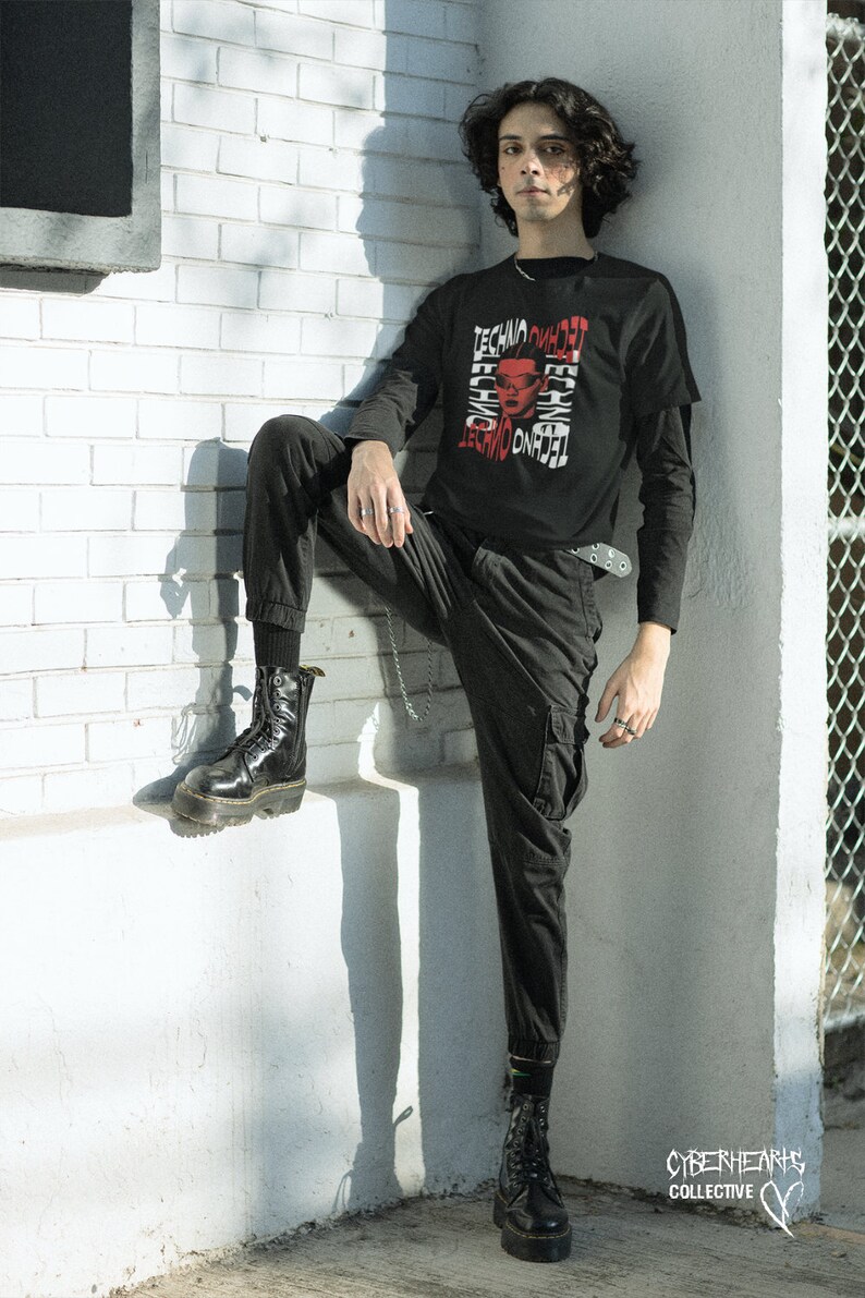 May include: A person wearing a black long-sleeved shirt with a red and white graphic print that says "Techno Omnia Vincit". The person is wearing black pants and black boots. The person is standing in front of a white brick wall.