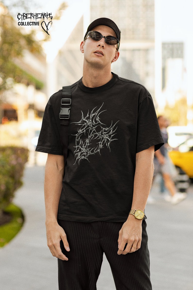 May include: A person wearing a black t-shirt with a white graphic design of a stylized heart. The graphic design has a barbed wire look. The person is wearing a black cap and sunglasses. The t-shirt has the text "CYBERHEARTS COLLECTIVE" on the top.