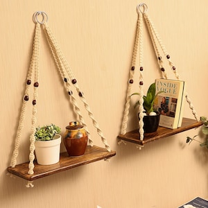 May include: Two wooden shelves with rope hangers and brown wooden beads. The shelves are hanging from the ceiling and are decorated with plants and a book titled "Inside Vogue".