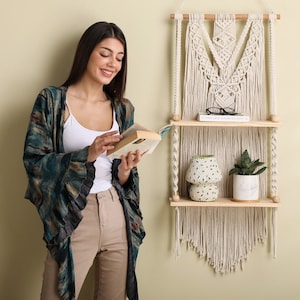 May include: A woman reading a book in front of a macrame wall hanging with two wooden shelves. The shelves are decorated with a potted plant, a decorative ceramic pot, and a pair of glasses. The wall hanging is cream colored and has a fringe.