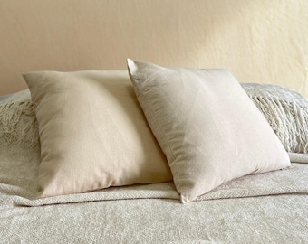 May include: Two beige square pillows rest on a bed covered with a textured blanket. The pillows are made of a soft material and have a simple, elegant design. The neutral color palette creates a calming atmosphere.