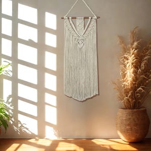 May include: Cream-colored macrame wall hanging with a geometric design and fringe. The wall decor is suspended from a wooden dowel and hangs against a neutral-toned wall. A woven basket with dried plants is in the background.