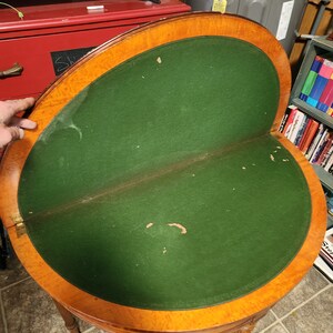 May include: A round wooden table with a green felt top. The table has a decorative wood trim around the edge and three legs.