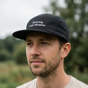 May include: A black five-panel cap featuring the white embroidered text Running Cult Member on the front crown. The hat has a flat brim, a structured front panel, and a small metal eyelet visible on the side panel for ventilation.