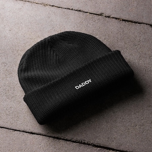 Peut inclure: Un bonnet en tricot noir uni avec un revers replié. Le mot DADDY est brodé en lettres capitales blanches au centre du revers. Le bonnet présente un motif en tricot côtelé texturé sur toute la surface du tissu.